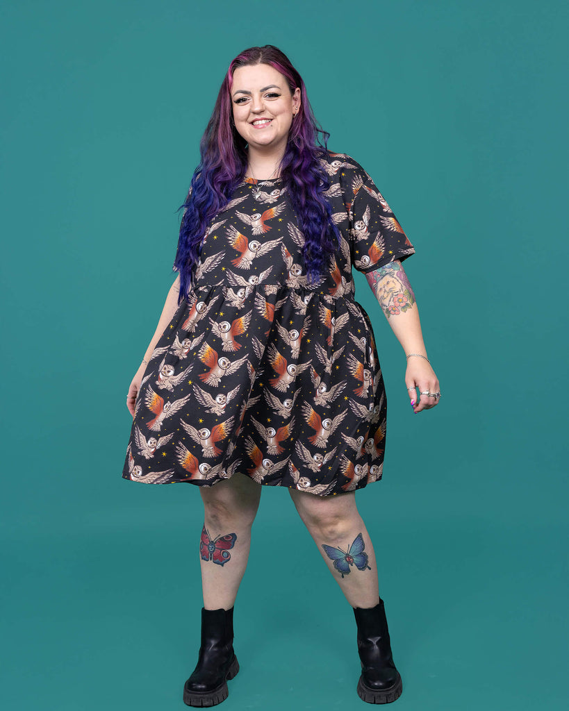 The What a Hoot Owl Oversized Smock Dress worn by a femme alternative tattooed model with long pink and purple hair, the matching tshirt and black boots on a teal studio background. She is facing forward smiling twirling in the dress with her arms out to the side. The black base dress features brown tawny owls flying amongst yellow stars.