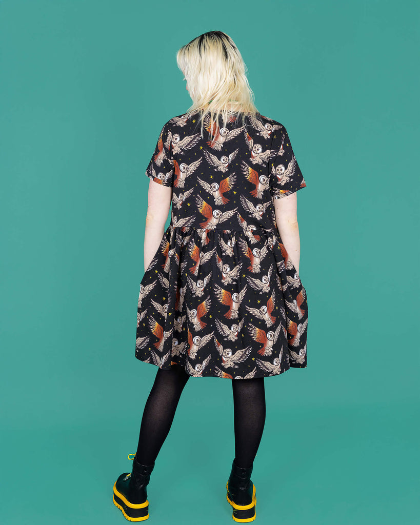 The What a Hoot Owl Oversized Smock Dress worn by a femme model with white blonde hair, black tights and yellow black boots on a teal studio background. She is facing away from camera with both hands resting in the dress pockets. The black base dress features brown tawny owls flying amongst yellow stars.