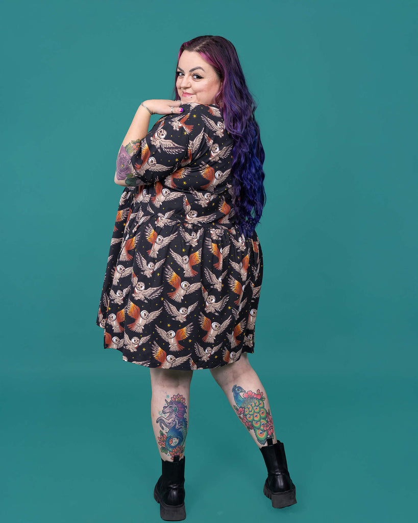 The What a Hoot Owl Oversized Smock Dress worn by a femme alternative tattooed model with long pink and purple hair, the matching tshirt and black boots on a teal studio background. She is facing away looking over her left shoulder to camera with her hand resting on her shoulder. The black base dress features brown tawny owls flying amongst yellow stars.