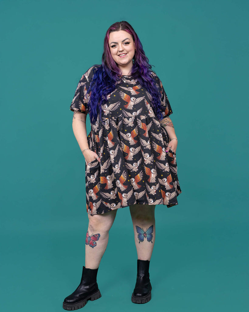 The What a Hoot Owl Oversized Smock Dress worn by a femme alternative tattooed model with long pink and purple hair, the matching tshirt and black boots on a teal studio background. She is facing forward smiling with both hands resting in the dress pockets. The black base dress features brown tawny owls flying amongst yellow stars.
