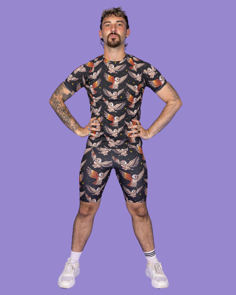 The What a Hoot Owl Short Sleeve T Shirt on a non binary masc presenting person with a short brown curly mullet haircut and the matching cycle shorts and white trainers on a purple studio background. They are facing forward with their hands on their hips pouting to camera. The black base tshirt features brown tawny owls flying amongst yellow stars.