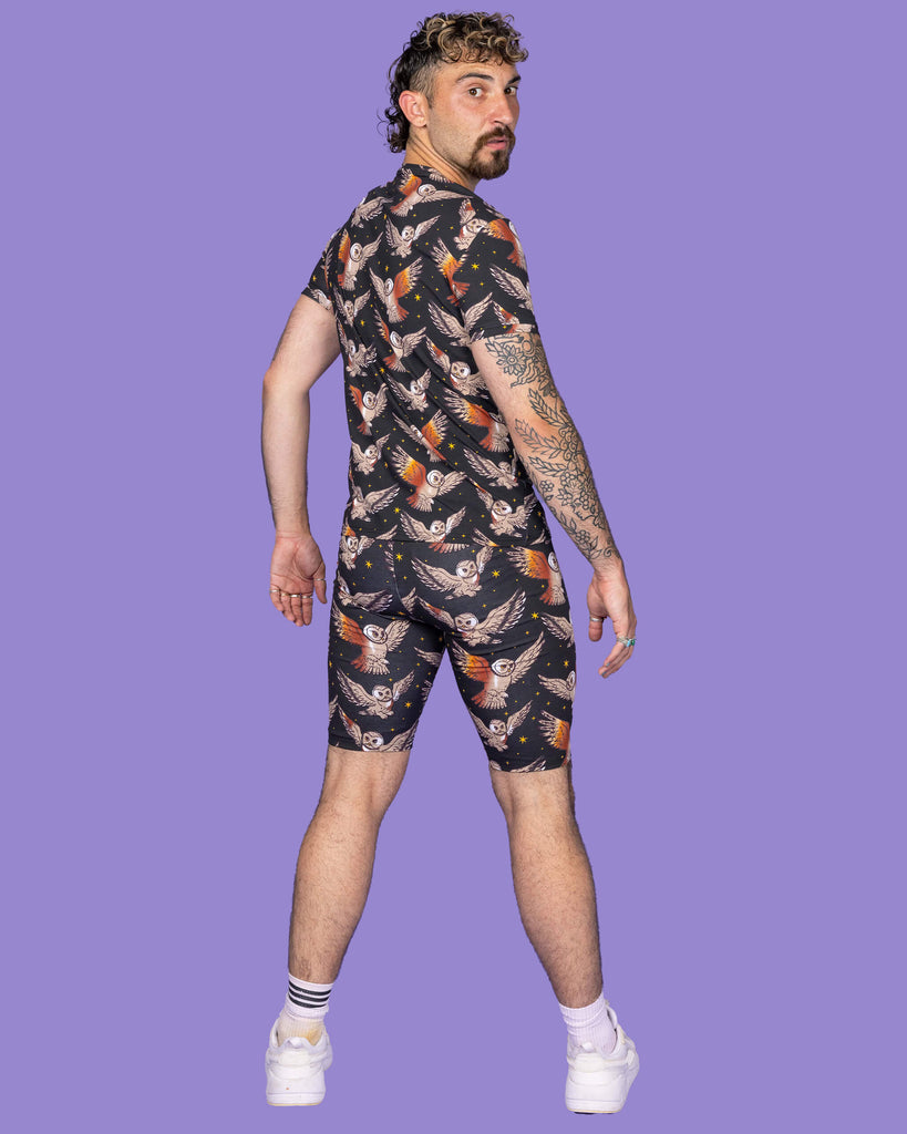 The What a Hoot Owl Short Sleeve T Shirt on a non binary masc presenting person with a short brown curly mullet haircut and the matching cycle shorts and white trainers on a purple studio background. They are facing away with their hands by their side looking over their right shoulder to camera. The black base t-shirt features brown tawny owls flying amongst yellow stars.