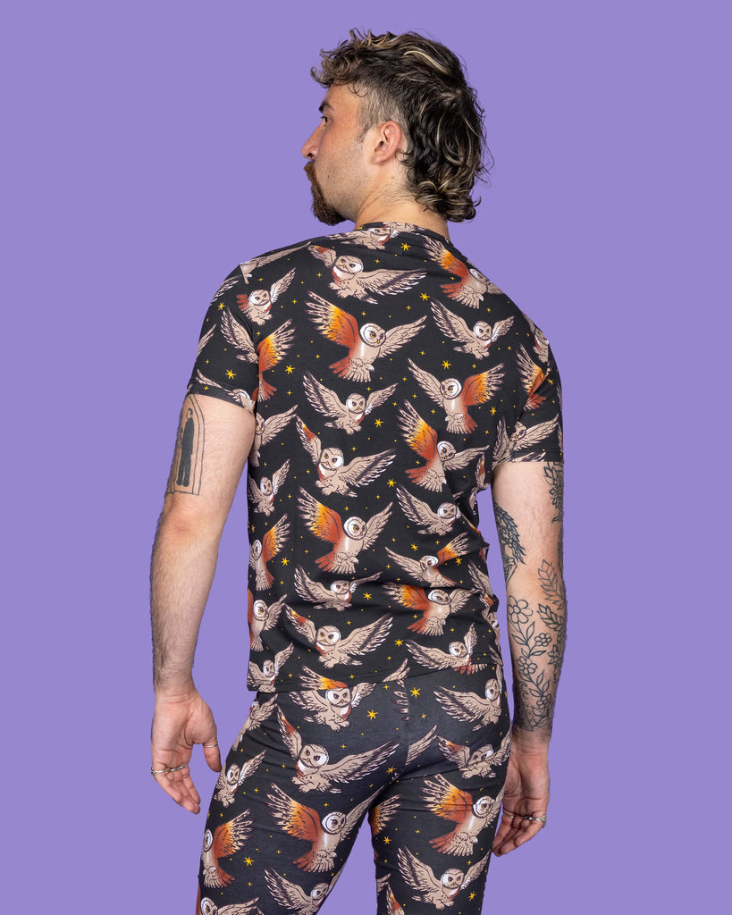 The What a Hoot Owl Short Sleeve T Shirt on a non binary masc presenting person with a short brown curly mullet haircut and the matching cycle shorts on a purple studio background. They are facing away with their arms by their side. The black base tshirt features brown tawny owls flying amongst yellow stars.