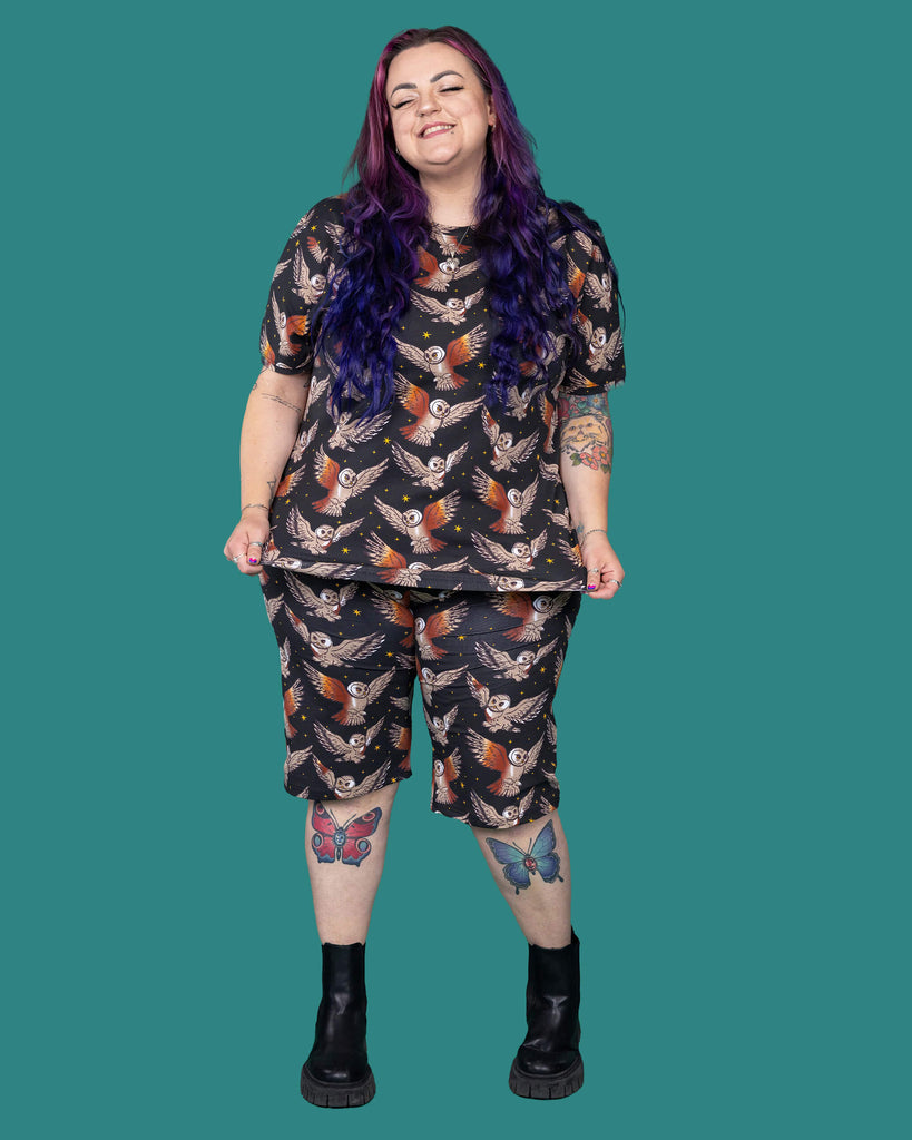 The What a Hoot Owl Short Sleeve T Shirt worn by a femme alternative tattooed model with long pink and purple hair, the matching cycle shorts and black boots on a teal studio background. She is facing forward laughing holding out the bottom of the tshirt. The black base tshirt features brown tawny owls flying amongst yellow stars.