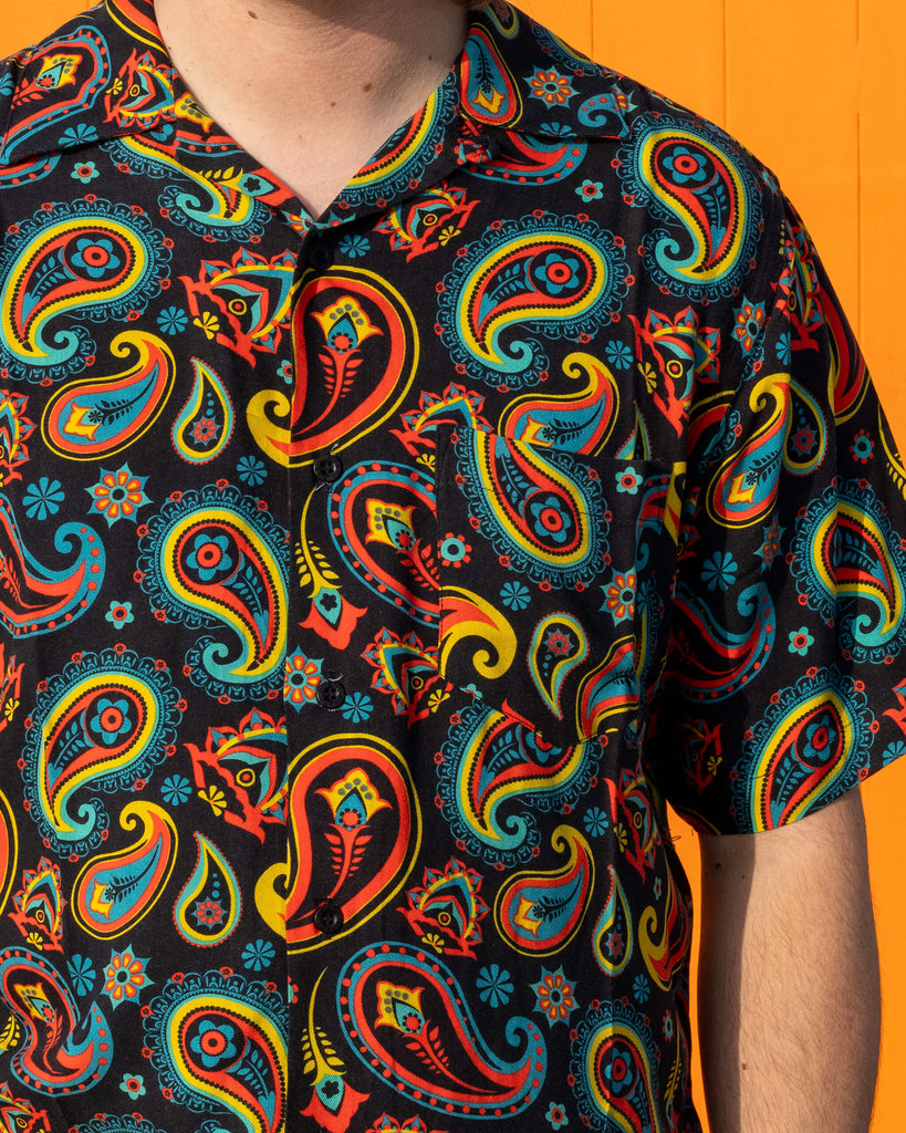 Close up of Michael wearing Run & Fly black paisley short sleeve shirt with dark blue jeans. The black shirt features an all over bold red, yellow, and blue paisley pattern. 