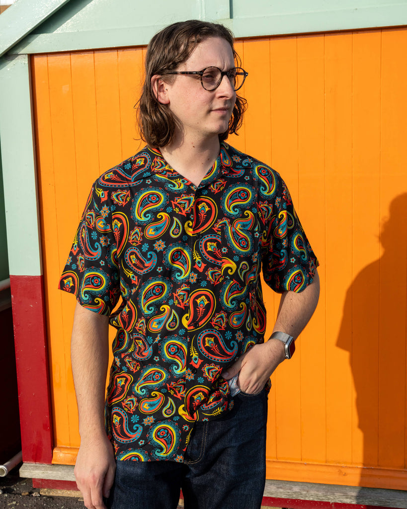 Michael is stood outside in Hove in front of beach huts wearing Run & Fly black paisley short sleeve shirt with dark blue jeans. The black shirt features an all over bold red, yellow, and blue paisley pattern. They are posing with one hand in their pocket and other down by their side looking off to one side.