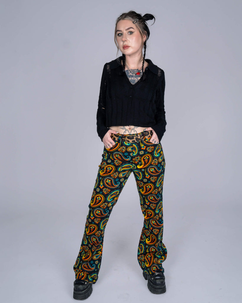  Model wearing black-and-colourful paisley print flared trousers paired with a cropped black knit top, striking a confident pose in a minimalist studio setting.