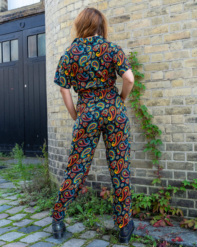 Back view of a model wearing a vibrant paisley jumpsuit by Run & Fly, an ethical slow fashion brand. The jumpsuit features colourful red, yellow, and blue patterns on a black background. Paired with black chunky boots, the look is stylish and bold. Run & Fly is known for their unique, sustainable designs that combine comfort with standout prints, perfect for those seeking conscious fashion choices.