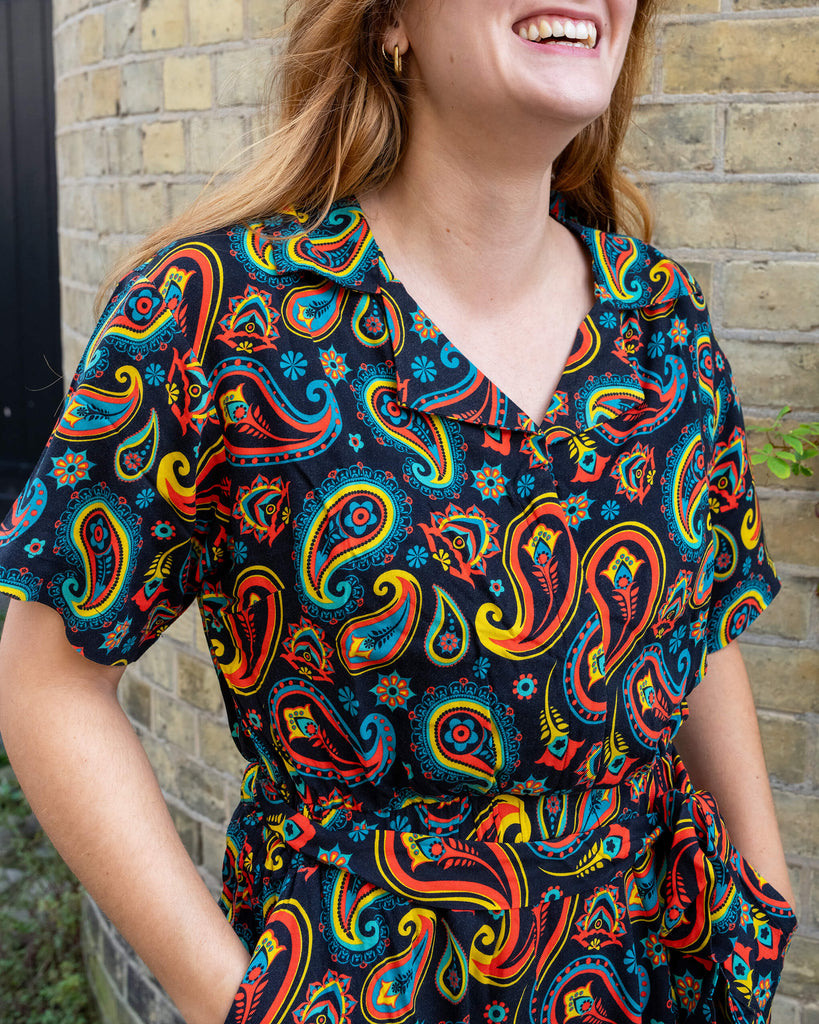Model laughing while wearing a colourful paisley jumpsuit by Run & Fly, an ethical slow fashion brand. The jumpsuit features bold red, yellow, and blue patterns on a black background. Run & Fly is renowned for their unique, sustainable fashion pieces that combine vibrant designs with comfortable style. Perfect for those seeking ethical, stand-out fashion choices.