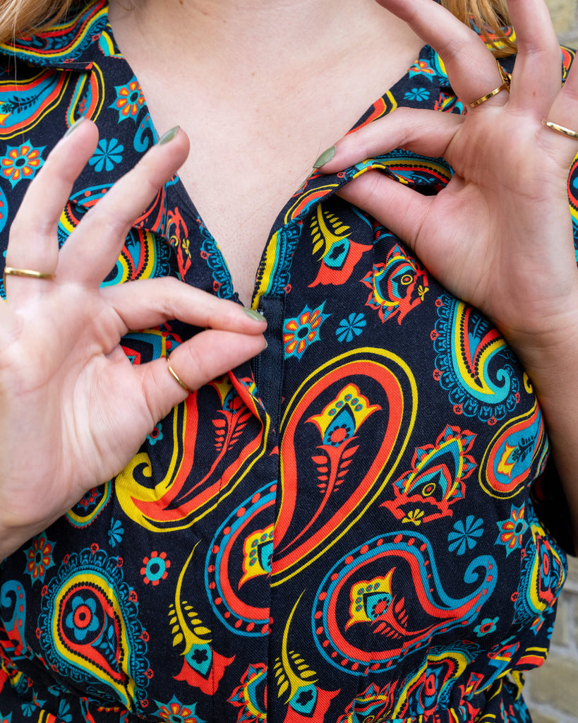 Close-up of a model adjusting the zip on a vibrant paisley jumpsuit from Run & Fly, an ethical slow fashion brand. The detailed pattern features bold red, yellow, and blue hues on a black background, highlighting the brand’s retro-inspired designs. The model's fingers showcase a delicate gold ring and green nail polish, adding a touch of elegance to the look. Run & Fly is committed to creating sustainable, high-quality fashion pieces that make a statement while supporting ethical practices.