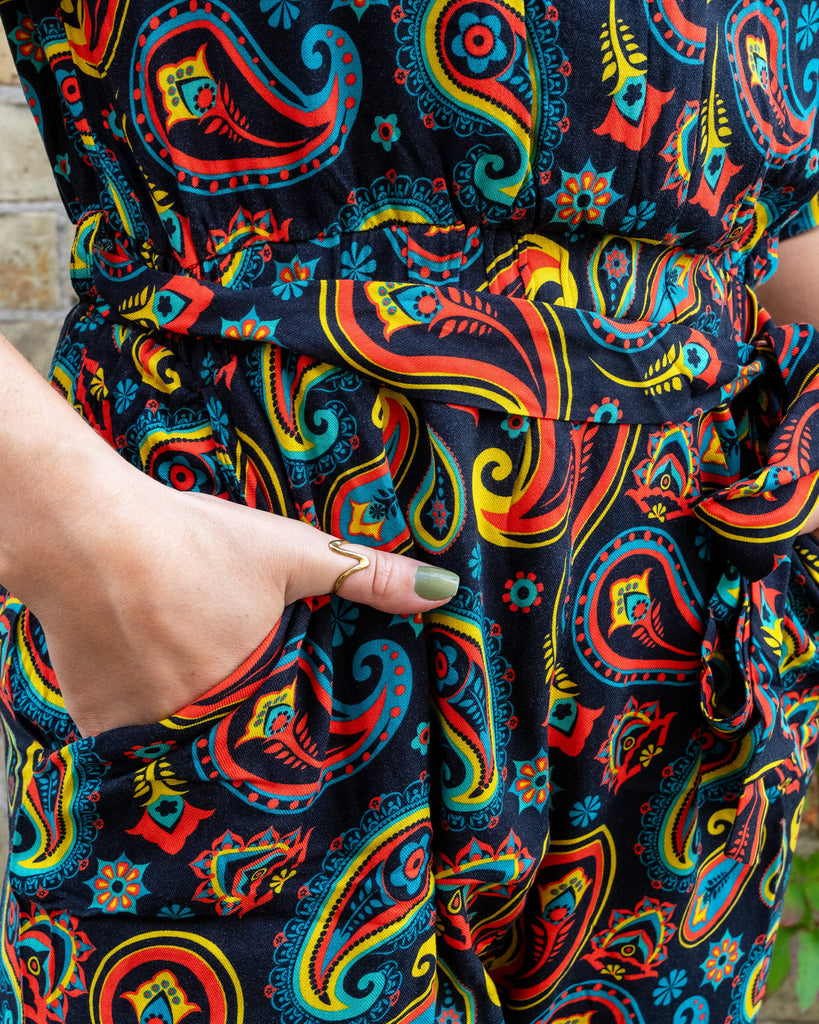 Close-up of a vibrant paisley jumpsuit from Run & Fly, an ethical slow fashion brand. The fabric features intricate red, yellow, and blue patterns on a black background, showcasing the brand's signature bold and retro-inspired designs. The model's hands are casually placed in the jumpsuit's pockets, highlighting its comfortable and practical style. Run & Fly is dedicated to creating sustainable, unique pieces that stand out for their quality and eye-catching prints, perfect conscious fashion lovers.