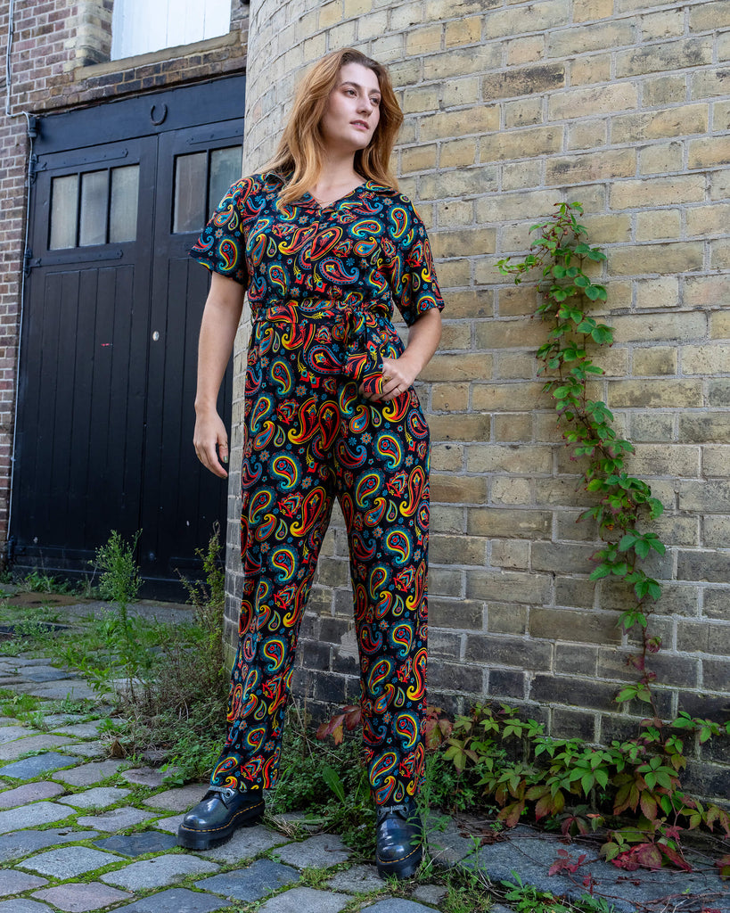 Model wearing a colourful paisley jumpsuit by Run & Fly, an ethical slow fashion brand, standing against a brick wall. The jumpsuit features bold red, yellow, and blue patterns, paired with black chunky boots. Run & Fly is known for its sustainable, retro-inspired designs and commitment to creating unique, high-quality fashion pieces.