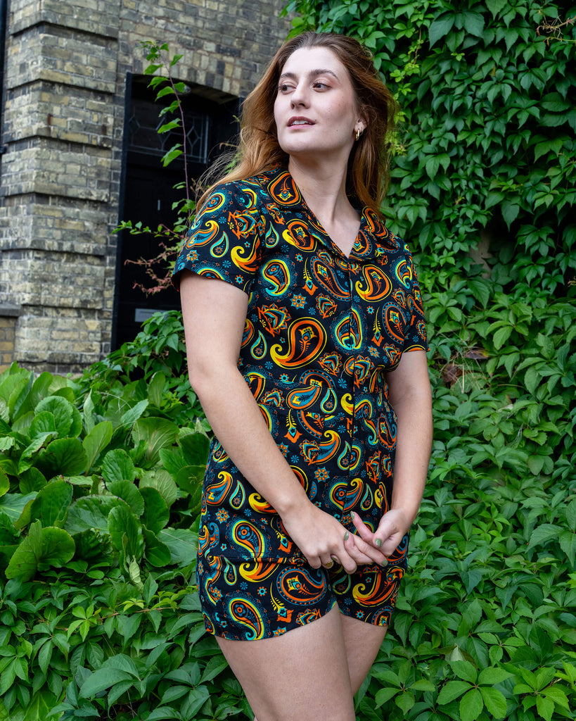 Model wearing a colourful paisley playsuit by Run & Fly, featuring vibrant orange, yellow, and blue patterns on a black background. The model stands against lush green foliage, showcasing the playsuit’s retro design. Run & Fly is known for its sustainable, ethically-made fashion pieces that blend bold prints with comfort and style.