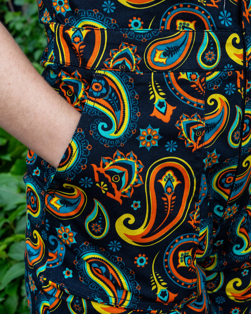 Close-up of a colourful paisley playsuit by Run & Fly, featuring bold orange, yellow, and blue patterns on a black background. The model’s hand is casually placed in the pocket, highlighting the playsuit’s practical and stylish design. Run & Fly is known for its vibrant, retro-inspired fashion and commitment to sustainable, ethical practices.