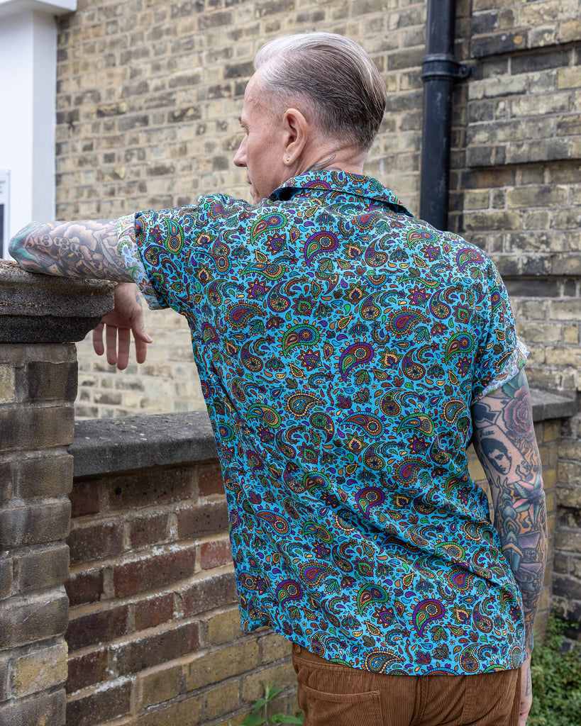 James is stood outside in a mews in Hove wearing wearing Sky Blue Paisley Short Sleeve Rayon Shirt paired with tan corduroy trousers. The collared, button up shirt is a sky blue colour with an all over paisley print in various muted rainbow colours. James is posing with his back to the camera and is leant sideways against a brick wall looking off to one side.