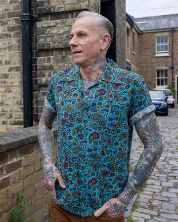 James is stood outside in a mews in Hove wearing wearing Sky Blue Paisley Short Sleeve Rayon Shirt paired with tan corduroy trousers. The collared, button up shirt is a sky blue colour with an all over paisley print in various muted rainbow colours. James is posing with his hands in his pockets and looking off to one side.