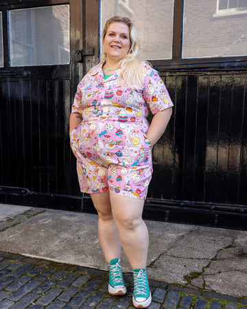 femme model with blonde hair is wearing Run & Fly x The Mushroom Babes Party Cats Stretch Playsuit with blue trainers. The collared playsuit is baby pink with white cats with party hats on and eating birthday cake, as well as various pink, blue and yellow sweets on the playsuit. The model is stood in a mews in Hove facing the camera with her hands in the pockets of the playsuit and smiling.