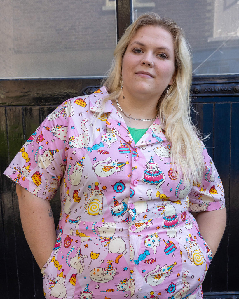femme model with blonde hair is wearing Run & Fly x The Mushroom Babes Party Cats Stretch Playsuit with blue trainers. The collared playsuit is baby pink with white cats with party hats on and eating birthday cake, as well as various pink, blue and yellow sweets on the playsuit. The model is stood in a mews in Hove facing the camera with her hands in the pockets of the playsuit and posing