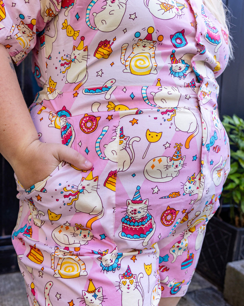 Close up of Party Stretch Playsuit. The playsuit is baby pink with white cats with party hats on and eating birthday cake, as well as various pink, blue and yellow sweets on the playsuit. The model's hand is in one of the pockets of the playsuit.