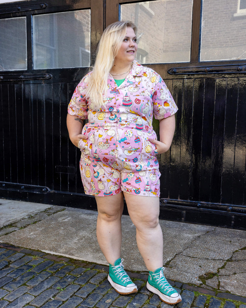 femme model with blonde hair is wearing Run & Fly x The Mushroom Babes Party Cats Stretch Playsuit with blue trainers. The collared playsuit is baby pink with white cats with party hats on and eating birthday cake, as well as various pink, blue and yellow sweets on the playsuit. The model is stood in a mews in Hove facing the camera with her hands in the pockets of the playsuit and looking off to one side