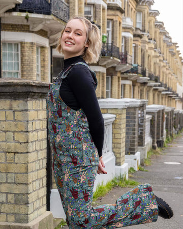 Model with short blonde hair wearing Peace and Love Stretch Skinny Flared Dungarees paired with long sleeve black turtleneck top and chunky black boots. The dungarees are an olive green colour with hands in different skin tones making the peace sign, various flowers, peace symbols and love hearts in white, yellow, purple and pink and 'peace' and 'love' printed on. There is a Run&Fly logo on front pocket on chest of dungarees and zip up side.