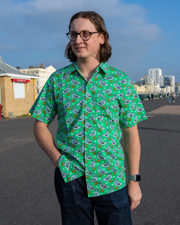 Michael is stood outside by Hove promenade wearing Run & Fly Pigeon Stars short sleeve shirt with dark blue jeans. The bright green shirt has an all over print of pigeons and white stars. Michael is posing looking off to one side with their hand in heir pocket. 