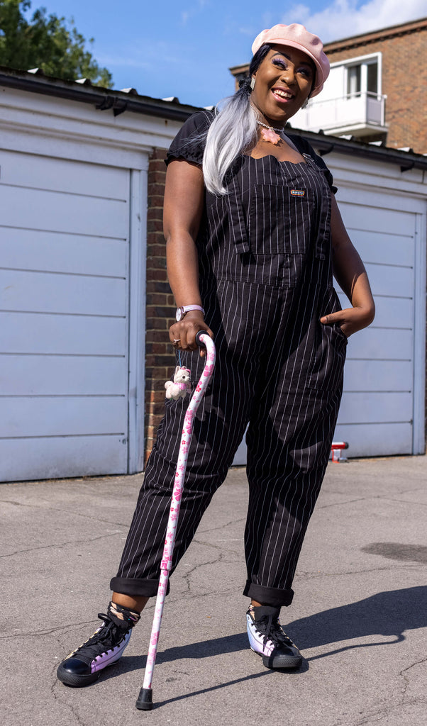 Ayesha a black model with black and white hair is smiling wearing black pinstripe dungarees and leaning on their mobility aid