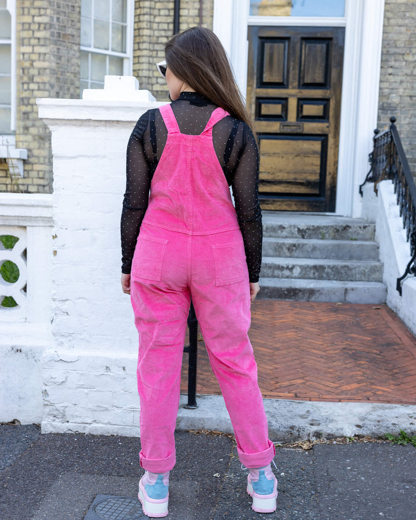 Charlotte is wearing Pink Stretch Corduroy Dungarees, paired with a long sleeved black mesh top, white framed sunglasses and chunky pink boots. The dungarees are a bright pink all over colour with Run&Fly logo on the front bib. Charlotte is posing with her back to the camera to show back of dungarees.
