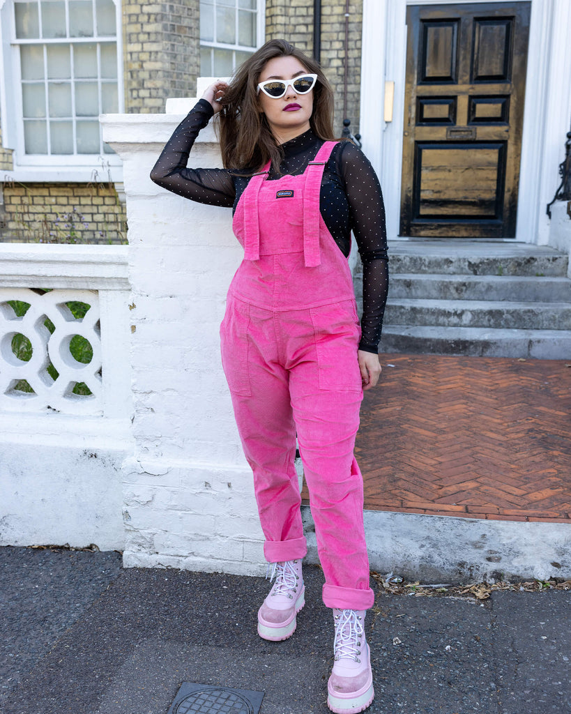 Charlotte is wearing Pink Stretch Corduroy Dungarees, paired with a long sleeved black mesh top, white framed sunglasses and chunky pink boots. The dungarees are a bright pink all over colour with Run&Fly logo on the front bib. Charlotte is posing with her hand in her hair and bottoms of the legs of dungarees rolled up to reveal her boots.