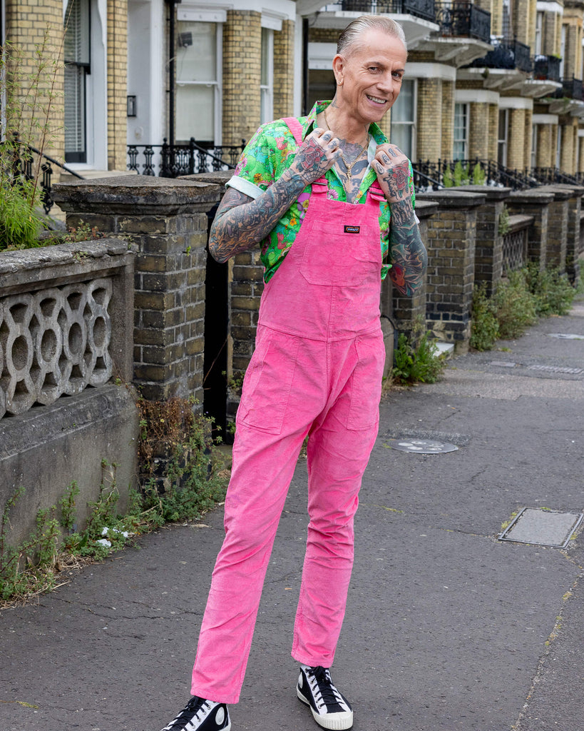 James is wearing Pink Stretch Corduroy Dungarees, paired with the Mushroom Babes frogs green short sleeve shirt and black and white trainers. The dungarees are a bright pink all over colour with Run & Fly logo on the front bib. James is posing with hands on collar camera to show front of dungarees.