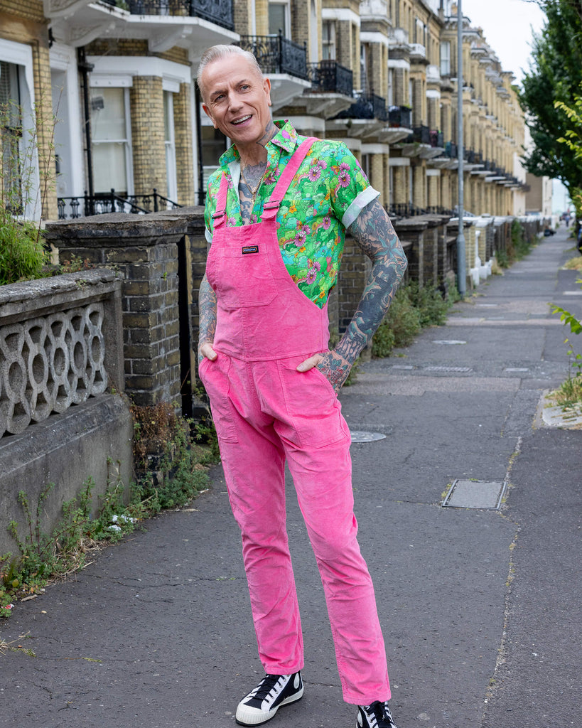 James is wearing Pink Stretch Corduroy Dungarees, paired with the Mushroom Babes frogs green short sleeve shirt and black and white trainers. The dungarees are a bright pink all over colour with Run & Fly logo on the front bib. James is posing with hands of the pockets of the dungarees.