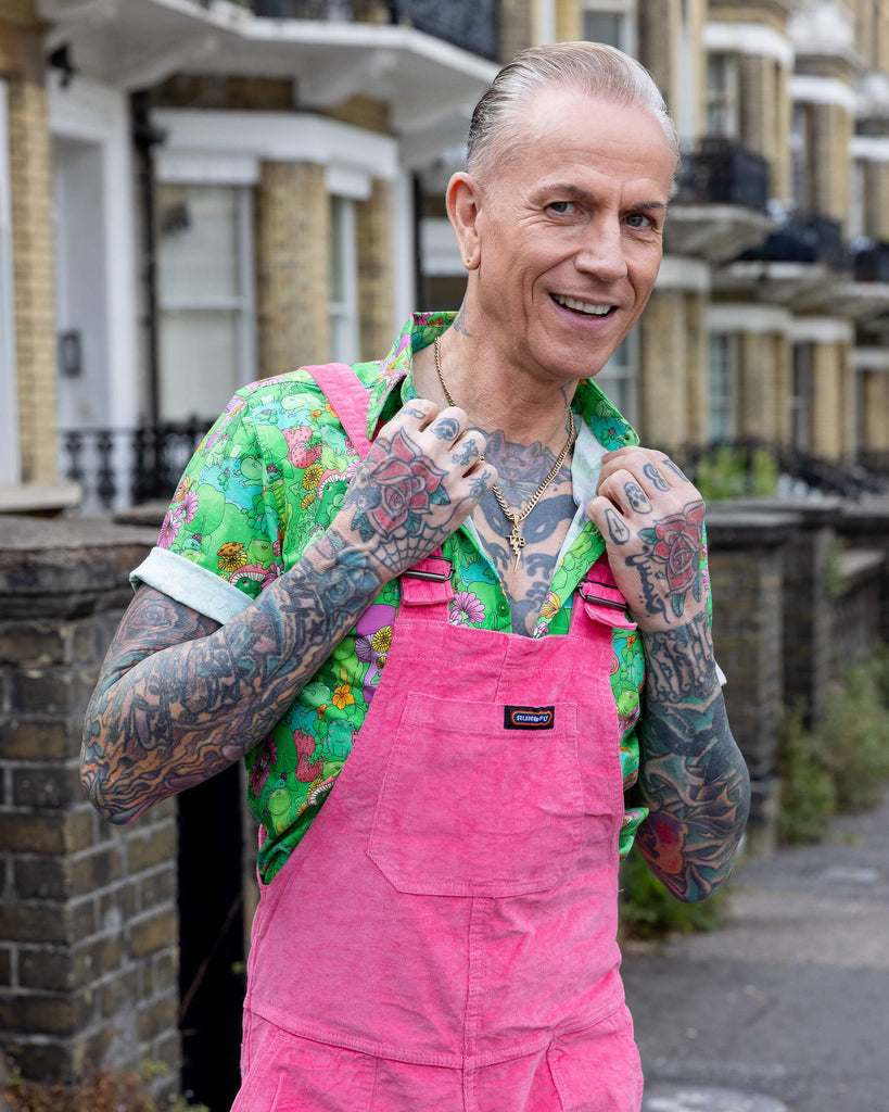 James is wearing Pink Stretch Corduroy Dungarees, paired with the Mushroom Babes frogs green short sleeve shirt and black and white trainers. The dungarees are a bright pink all over colour with Run & Fly logo on the front bib. James is posing with hands on collar camera to show front of dungarees.