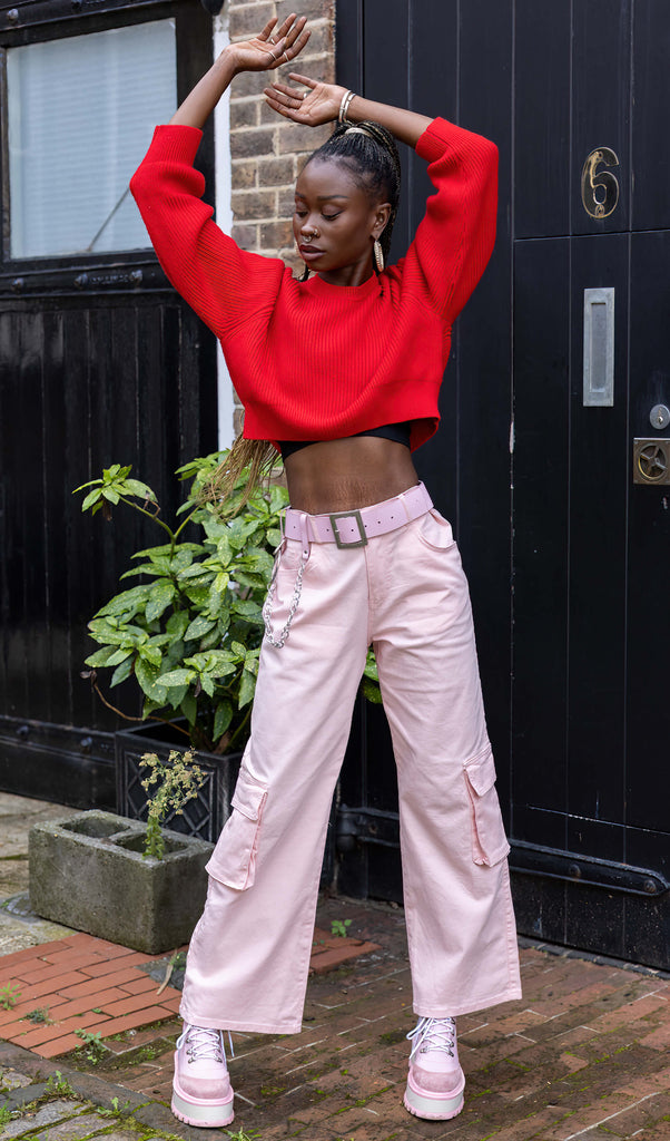 Yolande is stood outside in front of a black door wearing the 90's Baby Pink Wide Leg Oversized Stretch Cargo Trousers with a pink chain belt, red crop jumper and pink platform boots. She is facing forward leaning onto one hip with one hand in the side pocket, the other hand by her side, whilst looking off to the right. The trousers are an oversized fit, 2 flap cargo pockets on each leg as well as side pockets and back patch pockets she has her hands over her head and is looking to the side
