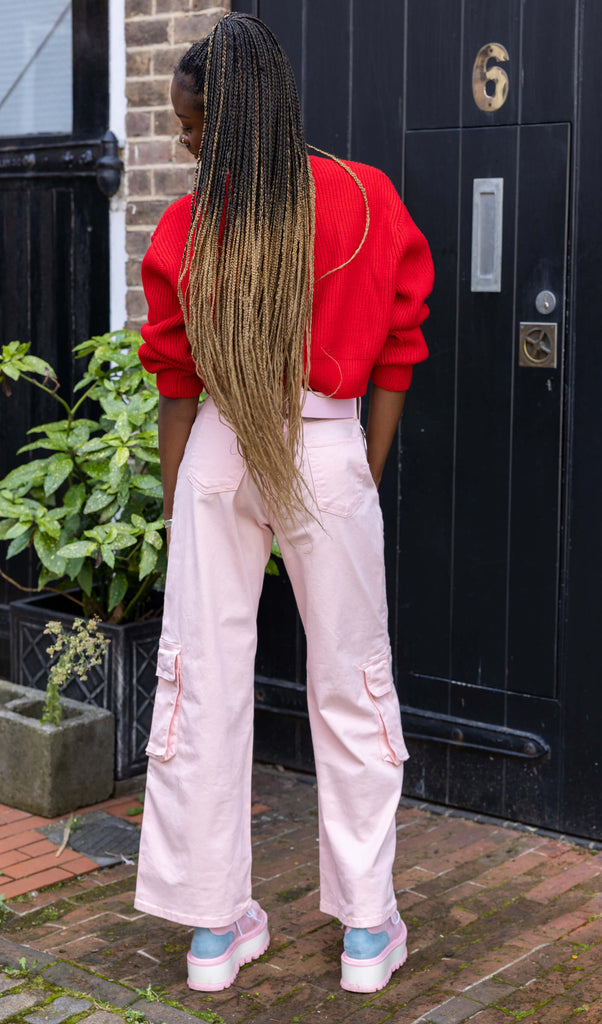 Yolande is stood outside in front of a black door wearing the 90's Baby Pink Wide Leg Oversized Stretch Cargo Trousers with a pink chain belt, red crop jumper and pink platform boots. She is facing forward leaning onto one hip with one hand in the side pocket, the other hand by her side, whilst looking off to the right. The trousers are an oversized fit, 2 flap cargo pockets on each leg as well as side pockets and back patch pockets.