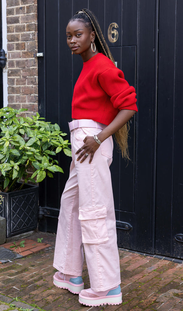 Yolande is stood outside in front of a black door wearing the 90's Baby Pink Wide Leg Oversized Stretch Cargo Trousers with a pink chain belt, red crop jumper and pink platform boots. She is facing forward leaning onto one hip with one hand in the side pocket, the other hand by her side, whilst looking off to the right. The trousers are an oversized fit, 2 flap cargo pockets on each leg as well as side pockets and back patch pockets.