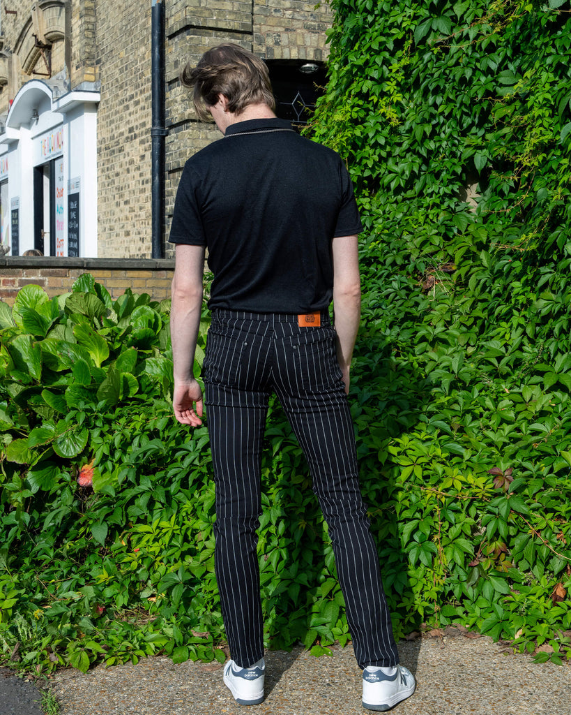 Jack, a white male model with light coloured hair is stood outside in Hove amongst greenery wearing Run & Fly Stretch Black & White Pinstriped Skinny Jeans with a short sleeved black tshirt tucked in and white trainers. He is posing with his back to the camera to show Run & Fly label on the back of the trousers.