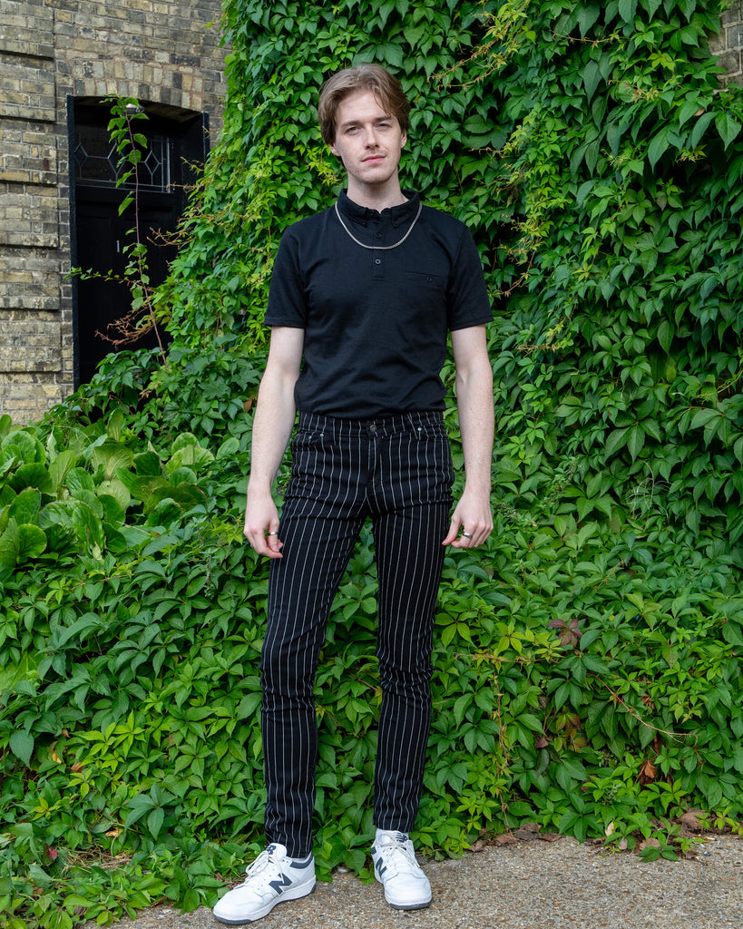 Jack, a white male model with light coloured hair is stood outside in Hove amongst greenery wearing Run & Fly Stretch Black & White Pinstriped Skinny Jeans with a short sleeved black tshirt tucked in and white trainers. He is posing facing the camera with his hands by his side. 