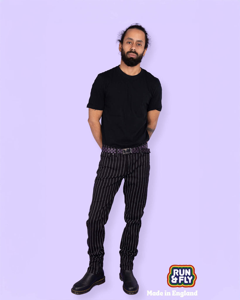Richard, a hispanic male model with dark hair in a bun and a beard, is stood in a photography studio in front of a lilac backdrop wearing Stretch Black & White Pinstriped Skinny Jeans with a black t shirt tucked in, a checkered belt and black boots. He is facing the camera with one leg slightly bent and his arms behind his back. Run & Fly logo is at the corner or the photo with 'Made in England' written underneath.