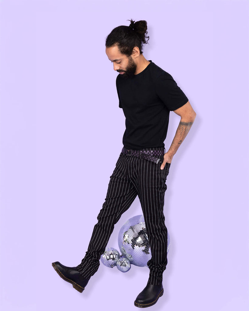 Richard, a hispanic male model with dark hair in a bun and a beard, is stood in a photography studio in front of a lilac backdrop amongst disco balls wearing Stretch Black & White Pinstriped Skinny Jeans with a black t shirt tucked in, a checkered belt and black boots. He is stood sideways with one leg stretched out in front of his and is looking down with his hands in the back pockets of the trousers.