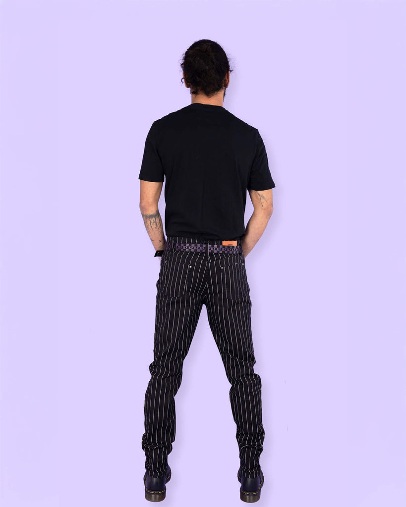 Richard, a hispanic male model with dark hair in a bun and a beard, is stood in a photography studio in front of a lilac backdrop amongst disco balls wearing Stretch Black & White Pinstriped Skinny Jeans with a black t shirt tucked in, a checkered belt and black boots. He is stood facing away with his hands in the side pockets of the trousers.
