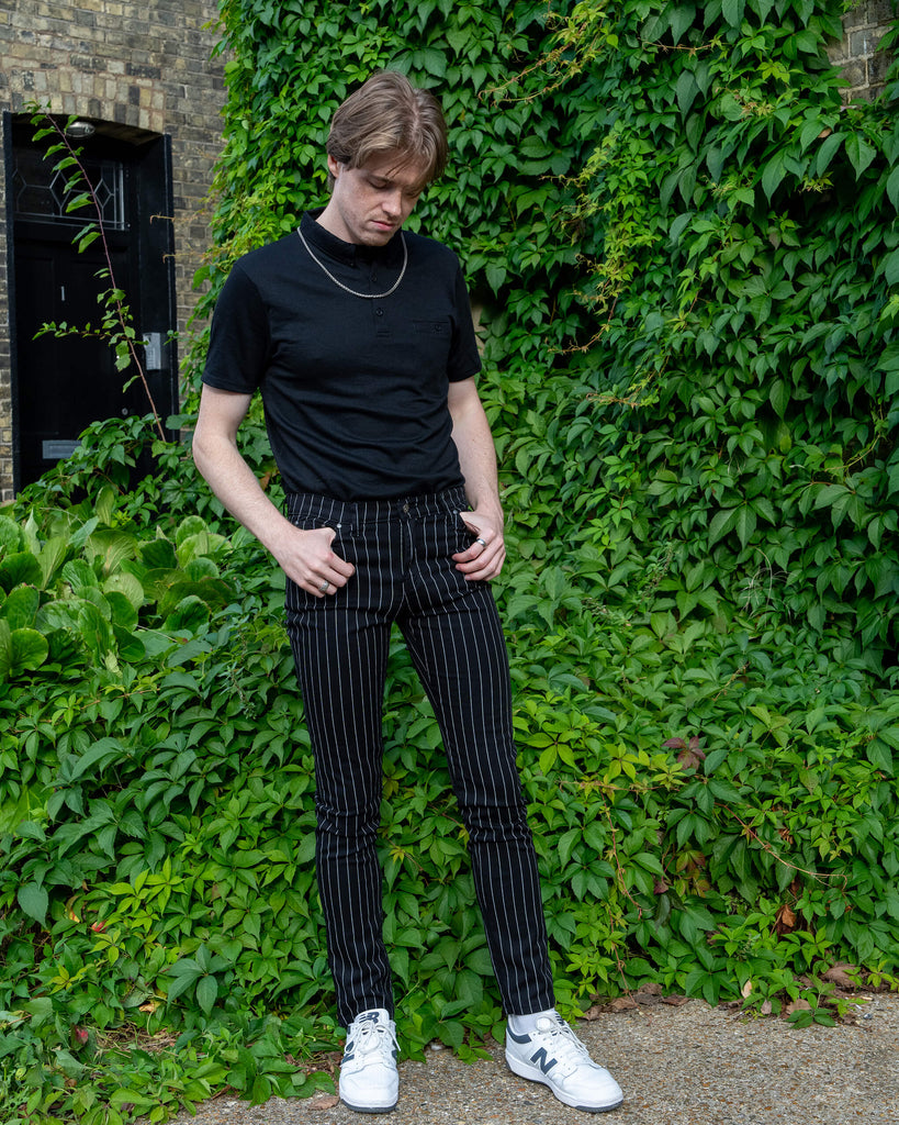 Jack, a white male model with light coloured hair is stood outside in Hove amongst greenery wearing Run & Fly Stretch Black & White Pinstriped Skinny Jeans with a short sleeved black tshirt tucked in and white trainers. He is facing the camera and looking down with his thumbs resting in the belt loops on the trousers. 