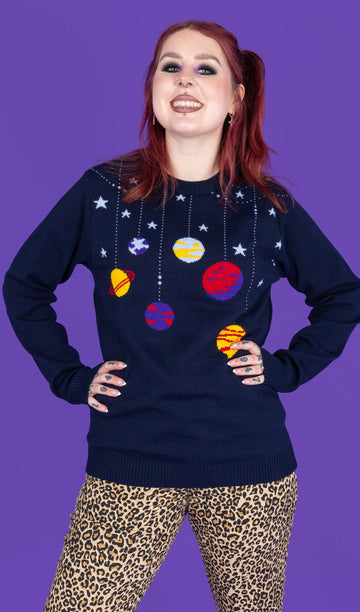 model with red hair and purple eye makeup wearing a navy coloured knit jumper with light blue stars and various planets on it, paired with leopard print jeans. The model is smiling at the camera with her hands on her hips. The background of the photo is purple.