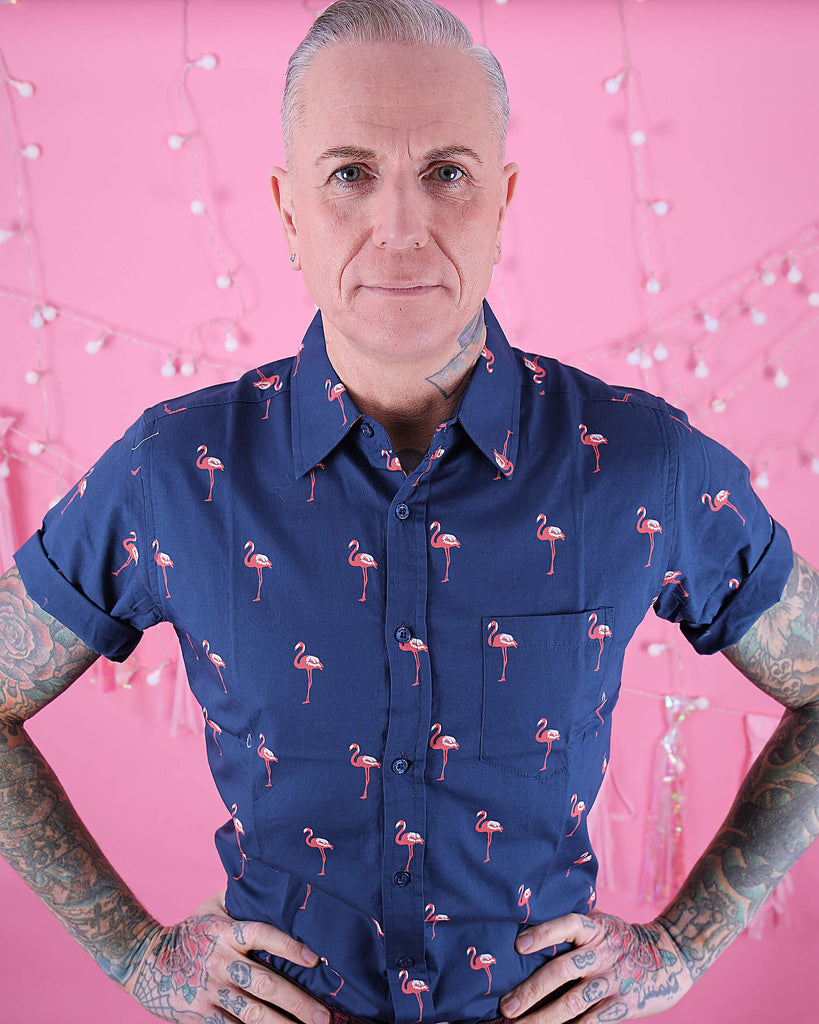 Jim is stood in front of a pink studio background wearing the preppy flamingo short sleeve shirt with burgundy trousers. They are heavily tattooed and have swept back silver hair. They are facing the camera and posing with both hands on their hips and smiling to the camera. Photo is cropped from the hips up.