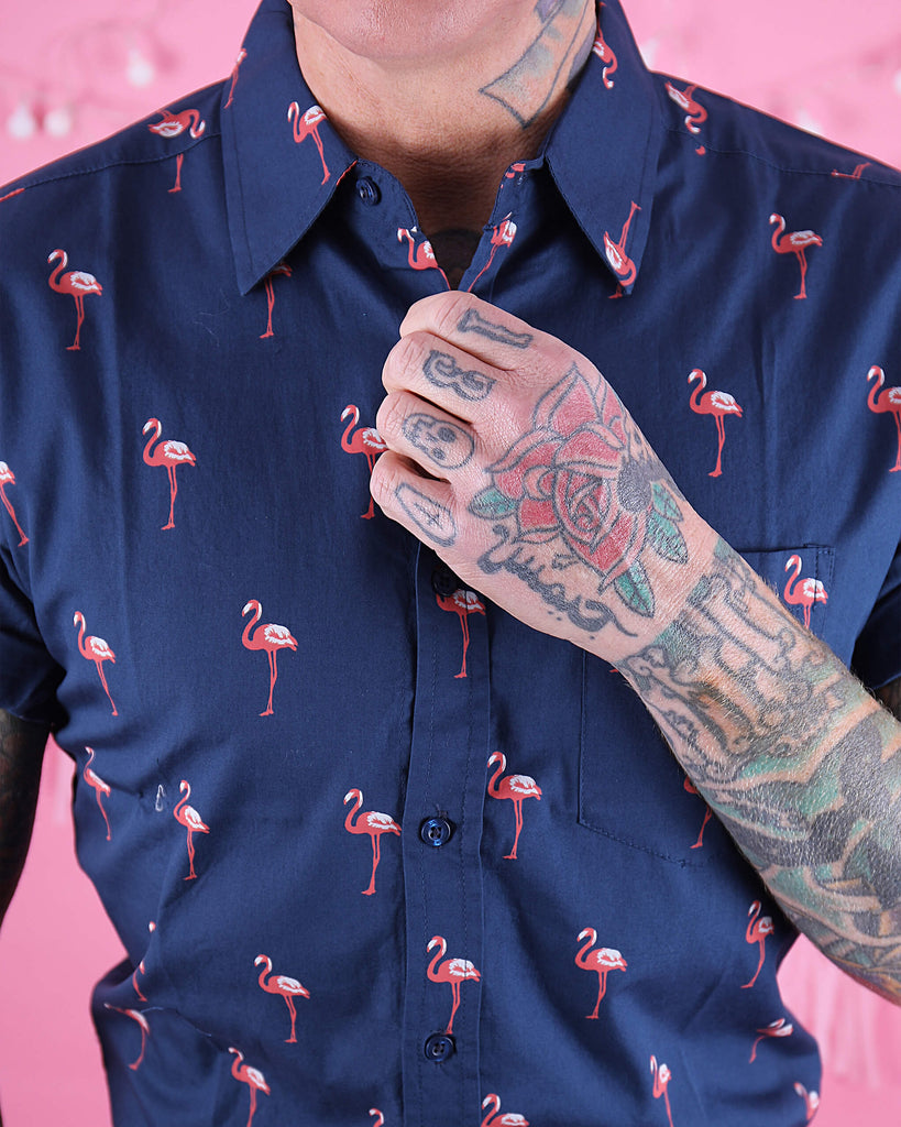 Close up shot of the front chest of the preppy flamingo short sleeve shirt. Heavily tattooed model has their hand going across doing up one of the top buttons.