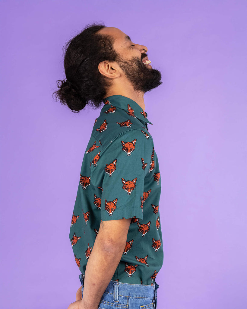 Richard, a hispanic male model with dark hair in a bun and a beard, is stood in a photography studio in Hove in front of a lilac backdrop wearing Preppy Foxy Short Sleeve Shirt with blue jeans. The emerald green shirt has an all over print of fox heads. Richard is stood sideways and is laughing with his eyes closed and head in the air. His hands are resting behind him and the photo is cropped at the thighs.