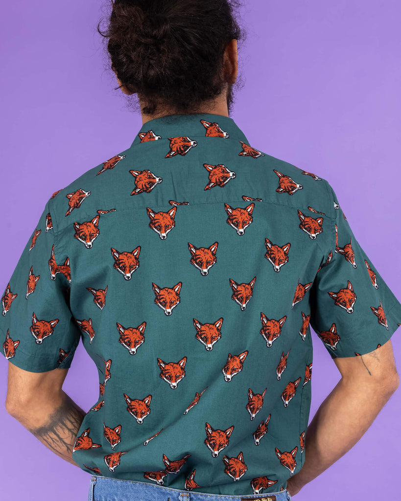 Richard, a hispanic male model with dark hair in a bun and a beard, is stood in a photography studio in Hove in front of a lilac backdrop wearing Preppy Foxy Short Sleeve Shirt with blue jeans. The emerald green shirt has an all over print of fox heads. Richard is posing with his back to the camera and his hands in front of him. The photo is cropped at the thighs.