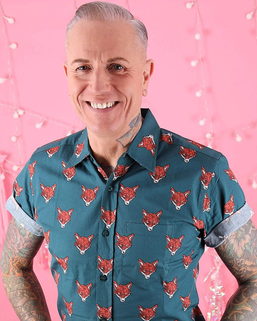 Jim is stood in front of a pink studio background wearing the preppy foxy short sleeve shirt tucked into brown corduroy trousers. They are heavily tattooed with swept back silver hair. They are facing the camera posing with both hands in their front trouser pockets laughing to camera. Photo is cropped from the hips up.