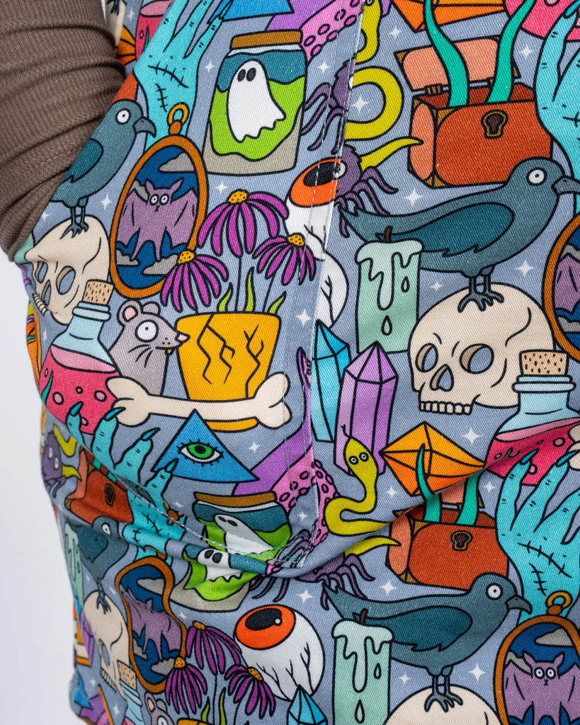 A close-up shot of the Pretty Spooky Pinafore highlights the intricate print, focusing on colourful details like potion bottles, ravens, spiders, skulls, and ghosts. The pocket detail is shown, emphasising its functionality and vibrant pattern.