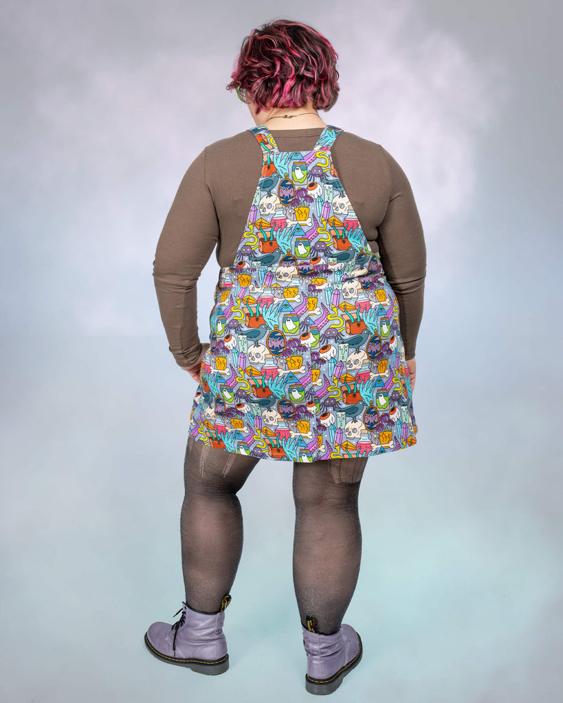 A back view of the Pretty Spooky Pinafore reveals the print extending across the back panel and straps. The grey background accentuates the colourful spooky illustrations like potions, mystical creatures, and other enchanting details. Jackie pairs the look with lilac Dr. Martens boots.