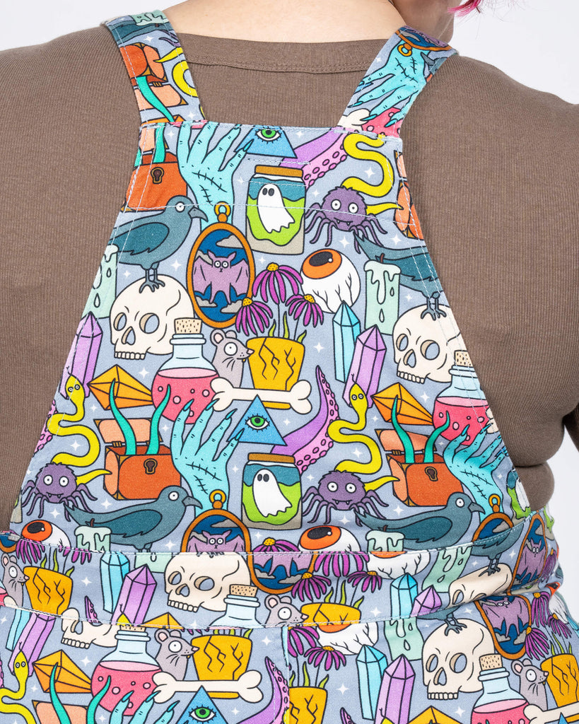 A close-up of the back panel highlights the intricate spooky illustrations and how the straps integrate seamlessly into the pinafore’s print. The grey background enhances the colourful design of skulls, ghosts, potions, and more.